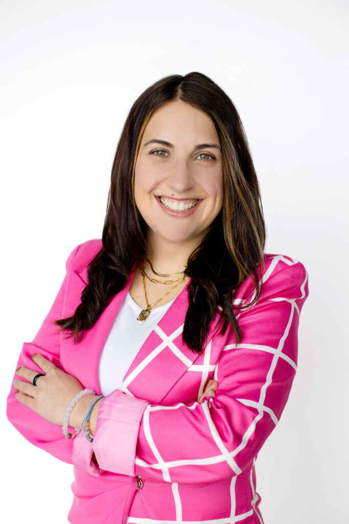 White woman with dark brown hair, wearing a pink patterned blazer.